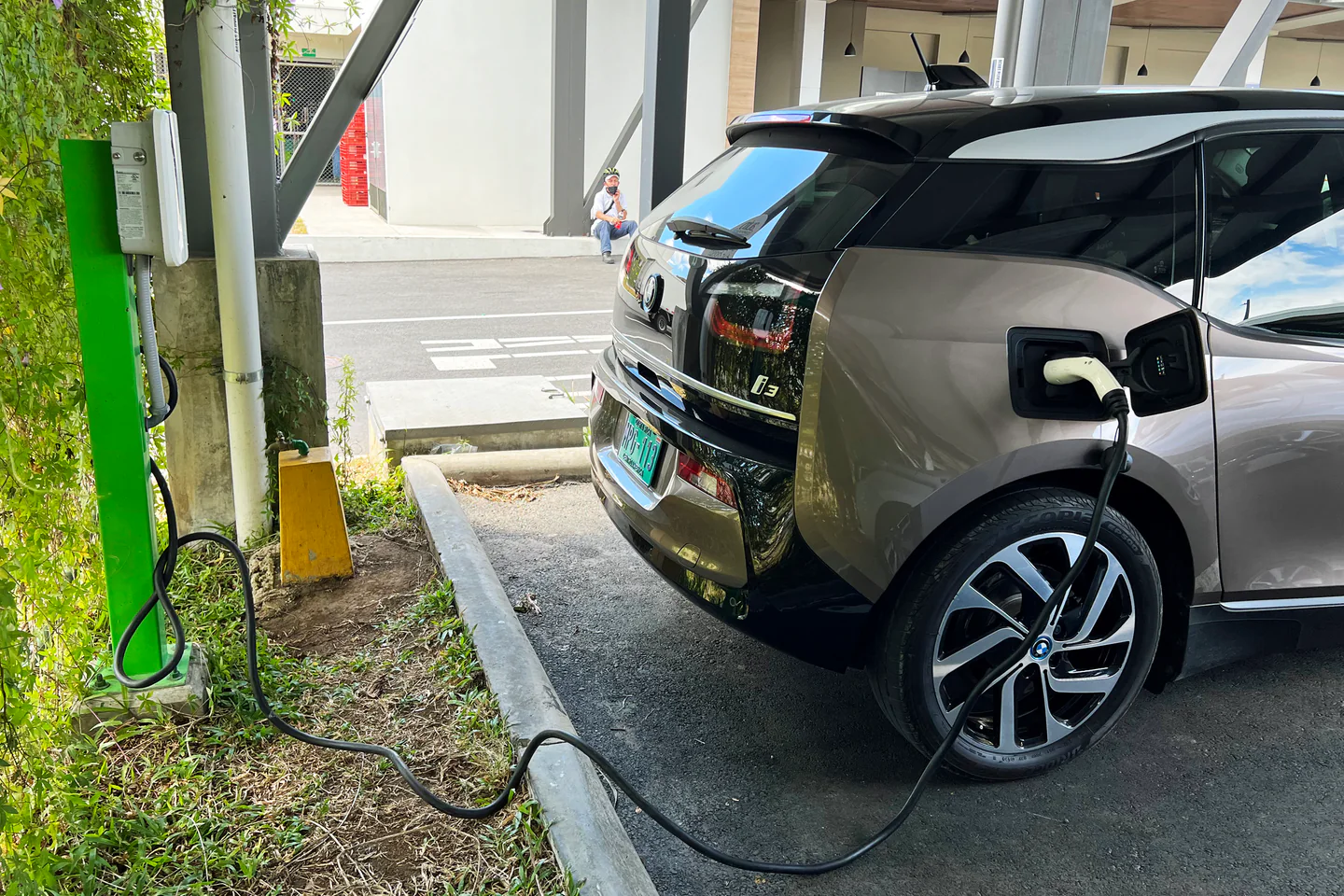 A white BMW i3 connected by cable to a public Level 2 charging station outside Automercado Guayabos in Costa Rica.